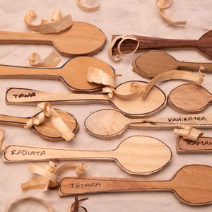 Könnte beinhalten: Eine Sammlung unfertiger Holzlöffel in verschiedenen hellen und dunklen Holztönen, jeweils beschriftet mit Namen wie Rimu, Kauri, Tawa, Cherry, Kahikatea, Kamahi, Radiata und Totara, begleitet von verstreuten Holzspänen auf hellem Stoff.