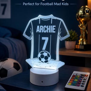 May include: Personalised football LED night light shaped like a football jersey, with the name "Archie" and the number 7. The light emits a bright white glow and sits on a white base. A remote control is included.
