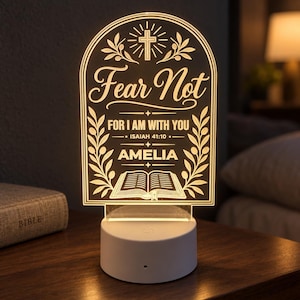May include: An arched, illuminated acrylic night light featuring a warm glow. The design includes a cross, olive branches, an open book, and the text: Fear Not, FOR I AM WITH YOU, ISAIAH 41:10, and the name AMELIA. It sits on a round white base.