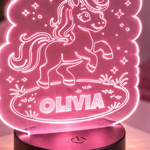 May include: A pink LED night light with a unicorn design and the name "OLIVIA". The light has a cloud-shaped outline with stars and a crescent moon. The base is black with a power symbol. The light is suitable for a child's bedroom.
