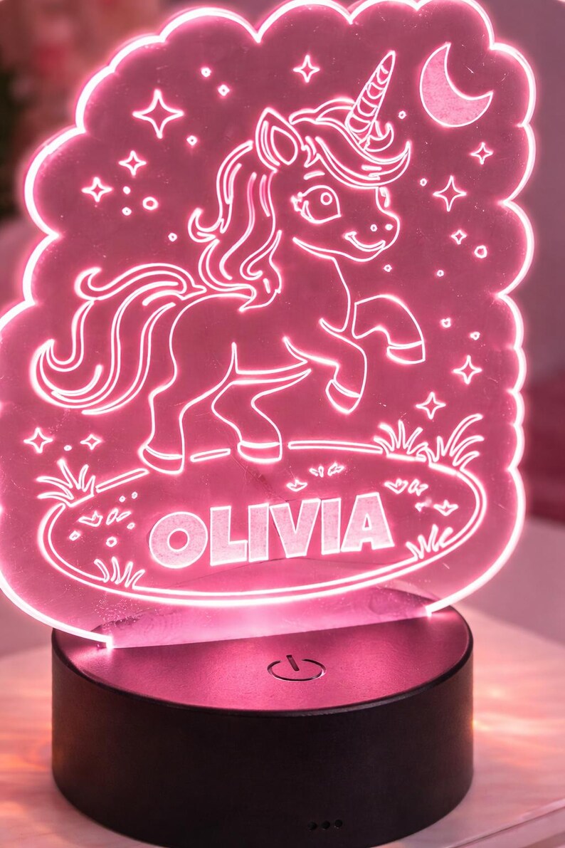 May include: A pink LED night light with a unicorn design and the name "OLIVIA". The light has a cloud-shaped outline with stars and a crescent moon. The base is black with a power symbol. The light is suitable for a child's bedroom.