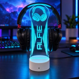 May include: A black headset rests on a clear acrylic stand with a white circular base. The stand features a glowing cyan light, an engraved headphone icon, and the name ALFIE in bold, vertical, capital letters illuminated in bright cyan.