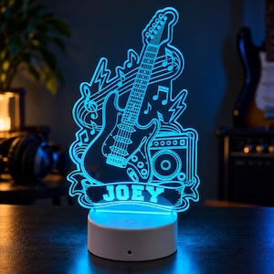 May include: A bright blue LED night light featuring an electric guitar, a musical amplifier, and musical notes. The design includes a banner at the bottom with the name JOEY printed in white block letters, all mounted on a circular white base.