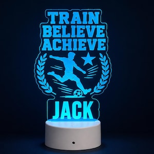 May include: A light blue LED acrylic lamp featuring a silhouette of a football player kicking a ball, surrounded by a laurel wreath and a star. The text reads TRAIN BELIEVE ACHIEVE and the name JACK, all illuminated against a dark background.