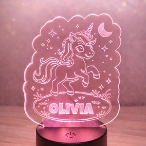May include: Personalised LED night light with a unicorn design. The light is pink and features the name "OLIVIA" in a decorative font. The unicorn is outlined in white and has a horn, mane, and tail. The base is black.