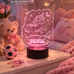 May include: A pink unicorn night light with the name "Olivia" illuminated. The light sits on a black base. The text "A magical personalised glow for her bedroom" is displayed. Ideal for a child's bedroom.