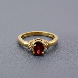 Op de afbeelding: Een gouden ring met een grote, ovaal geslepen, dieprode edelsteen als middelpunt. De gepolijste gouden band heeft kleine accentstenen aan weerszijden van de hoofdsteen. De ring is geplaatst tegen een grijze achtergrond.