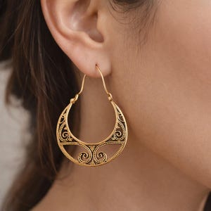 Puede incluir: Pendientes de aro dorados con un intrincado diseño de filigrana. Los pendientes tienen forma de media luna con volutas detalladas y patrones de puntos. Los pendientes se muestran en una oreja.
