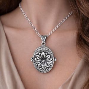 Könnte beinhalten: Silberfarbene ovale Medaillon-Halskette mit floralem Design. Der Anhänger zeigt eine geprägte Blume und dekorative Details. Die Halskette hat eine zarte Kette und wird um den Hals getragen.