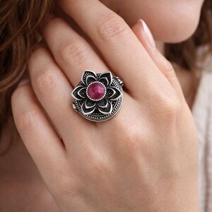 May include: A silver-toned ring featuring a flower design with a central, round, red gemstone. The ring's petals are detailed with etched lines, and the band has a textured pattern. The ring is worn on a finger.