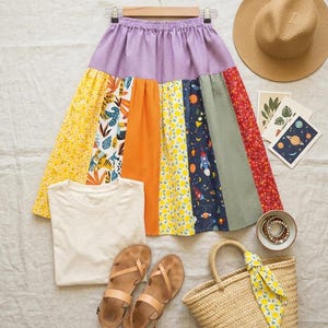 Puede incluir: Una colorida falda patchwork con cintura de lunares morados, combinada con una camiseta blanca, sandalias, un sombrero de paja y un bolso tote tejido. La falda presenta paneles con varios diseños, incluyendo florales, cítricos y temáticos espaciales. Pulseras y postales.