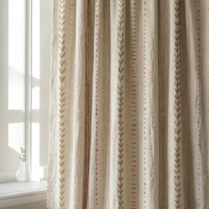 May include: A beige curtain panel featuring vertical stripes with repeating leaf patterns in olive green and small dotted lines in terracotta red. The fabric has a visible woven texture and hangs in soft, vertical folds, partially covering a window.