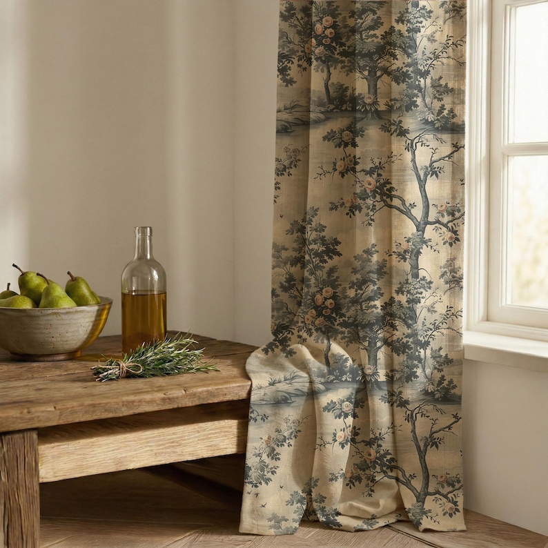 May include: A floor-length curtain featuring a dark blue and beige toile pattern of trees and flowers. Beside the curtain sits a clear glass bottle filled with golden liquid and a gray bowl containing several green pears and a sprig of rosemary.