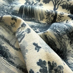 May include: A piece of cream-colored fabric featuring a dark blue toile de jouy print. The pattern depicts figures in period clothing standing in a landscape with trees and foliage. The textile has a visible woven texture and is folded to show depth.