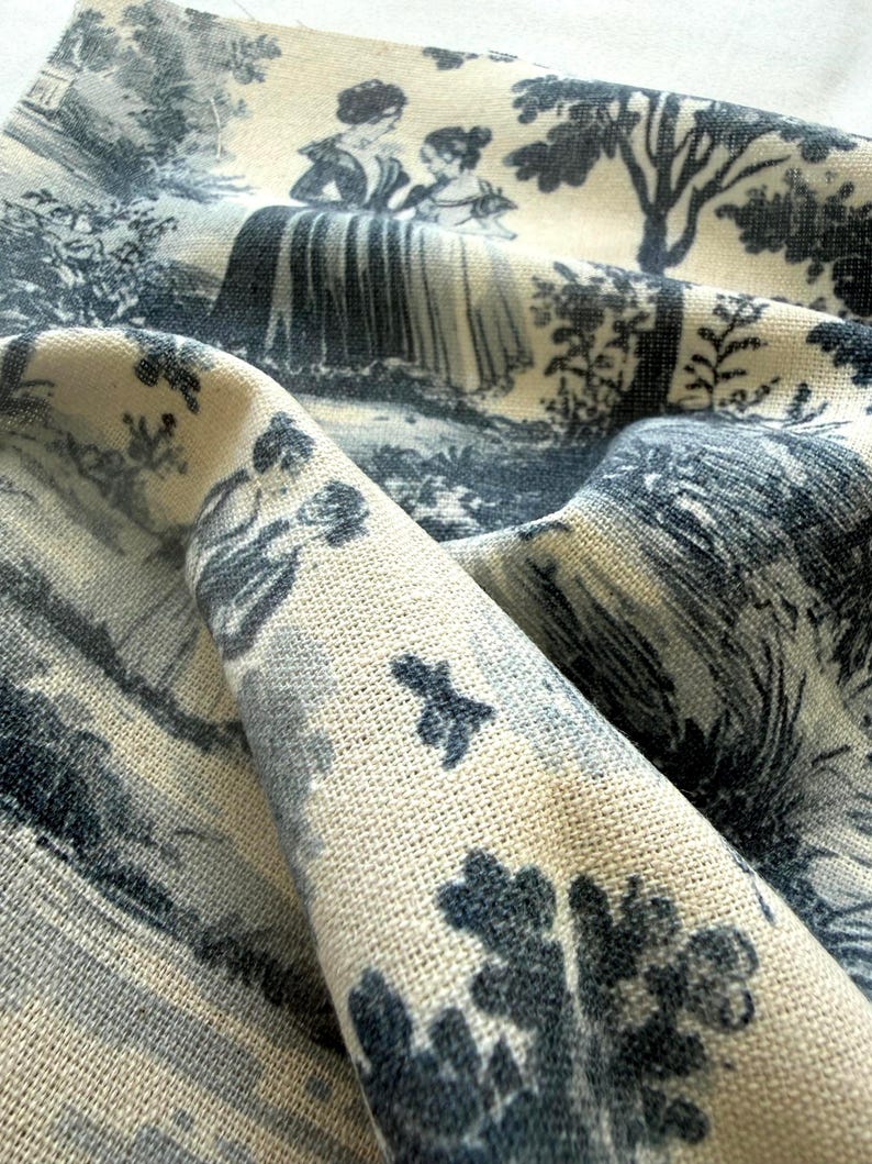 May include: A piece of cream-colored fabric featuring a dark blue toile de jouy print. The pattern depicts figures in period clothing standing in a landscape with trees and foliage. The textile has a visible woven texture and is folded to show depth.