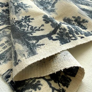 May include: A piece of off-white fabric featuring a dark blue toile de jouy pattern. The design displays detailed illustrations of trees, foliage, and architectural structures printed across the textured, woven surface of the textile material.