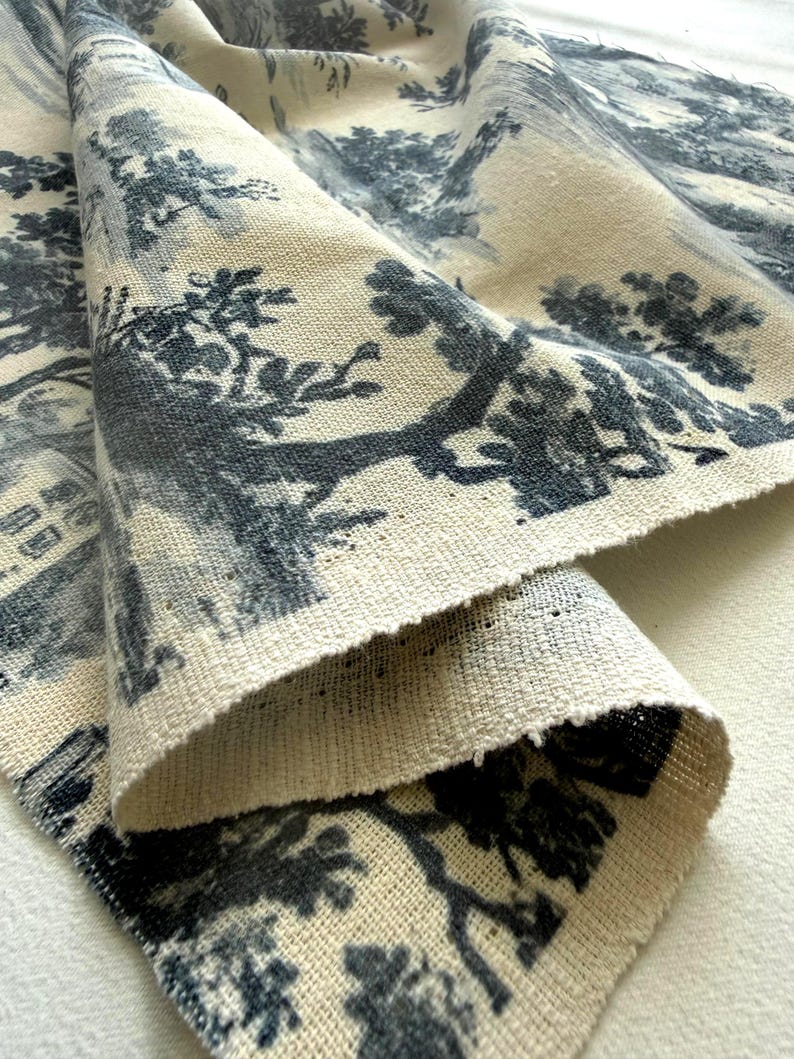 May include: A piece of off-white fabric featuring a dark blue toile de jouy pattern. The design displays detailed illustrations of trees, foliage, and architectural structures printed across the textured, woven surface of the textile material.