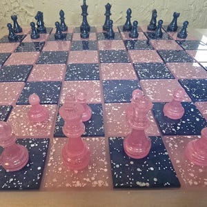 May include: A decorative chess set with a pink and navy blue speckled board. The chess pieces are translucent pink and navy blue. The board is divided into alternating pink and navy blue squares.