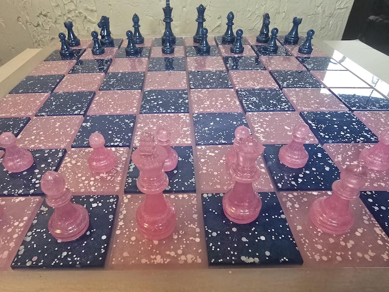 May include: A decorative chess set with a pink and navy blue speckled board. The chess pieces are translucent pink and navy blue. The board is divided into alternating pink and navy blue squares.