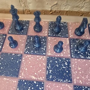 May include: A close-up view of a chess set with dark blue pieces on a board with pink and dark blue speckled squares. The pieces include a king, queen, bishop, knight, rook, and pawns. The board is set up for a game.