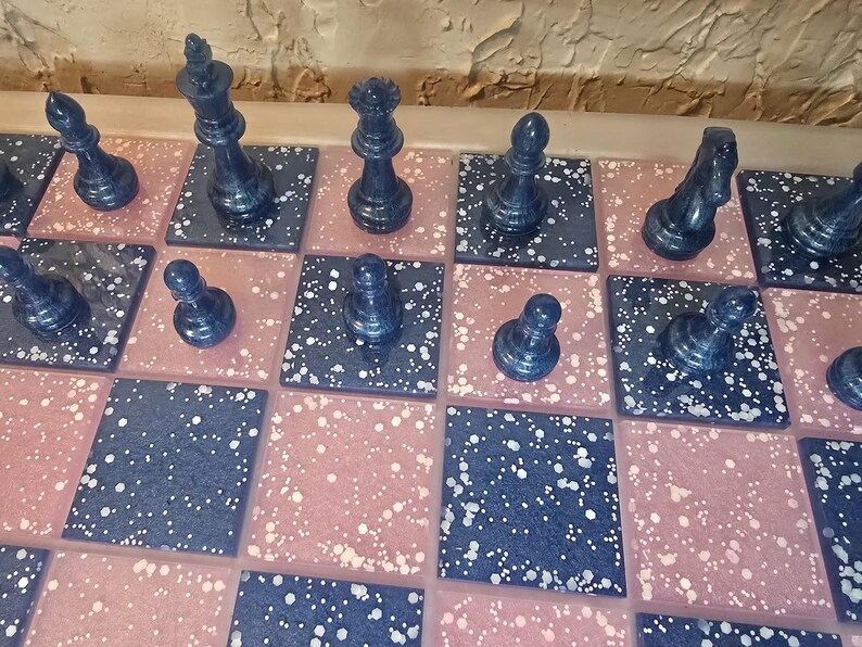May include: A close-up view of a chess set with dark blue pieces on a board with pink and dark blue speckled squares. The pieces include a king, queen, bishop, knight, rook, and pawns. The board is set up for a game.