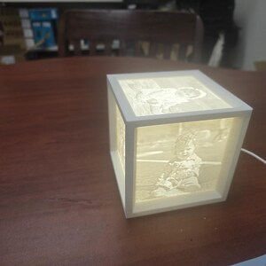 May include: A white, illuminated cube-shaped lamp with images on each side. The images appear to be of a baby. The lamp is sitting on a dark brown wooden surface. A white cord extends from the lamp.