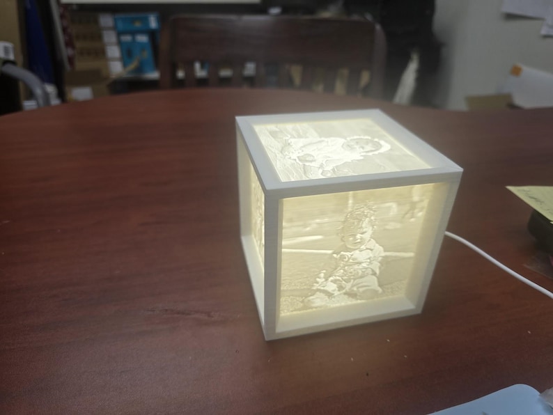 May include: A white, illuminated cube-shaped lamp with images on each side. The images appear to be of a baby. The lamp is sitting on a dark brown wooden surface. A white cord extends from the lamp.