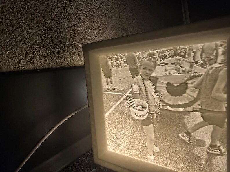 May include: A framed black and white photograph of a child at a parade. The child is holding a small basket with the word "Luther" on it and an American flag. The child is wearing a beaded necklace.