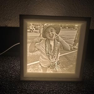May include: A sepia-toned, illuminated picture frame displaying a photograph of a person making a playful gesture. The person is wearing a hat, beads, and shorts. The frame has a soft, glowing light.