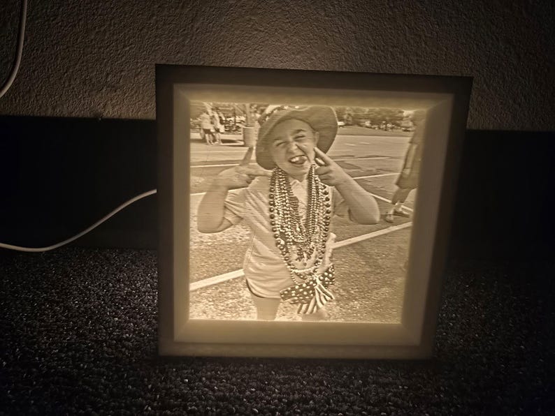 May include: A sepia-toned, illuminated picture frame displaying a photograph of a person making a playful gesture. The person is wearing a hat, beads, and shorts. The frame has a soft, glowing light.