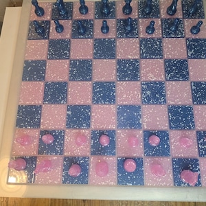 May include: A handmade chess set with a pink and blue checkered board. The board has a speckled design. The chess pieces are blue and pink. The set is displayed on a white surface.