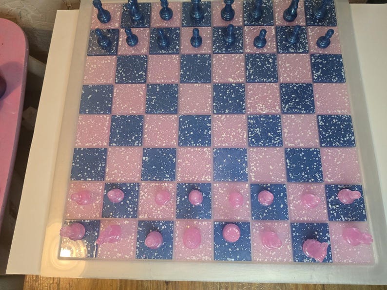 May include: A handmade chess set with a pink and blue checkered board. The board has a speckled design. The chess pieces are blue and pink. The set is displayed on a white surface.