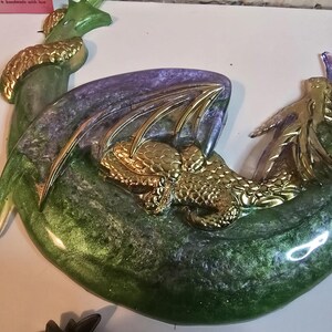 May include: A decorative dragon sculpture in shades of green, purple, and gold. The dragon is curled up, with gold-colored wings and accents. The base has a crescent moon shape. The sculpture is made of resin.