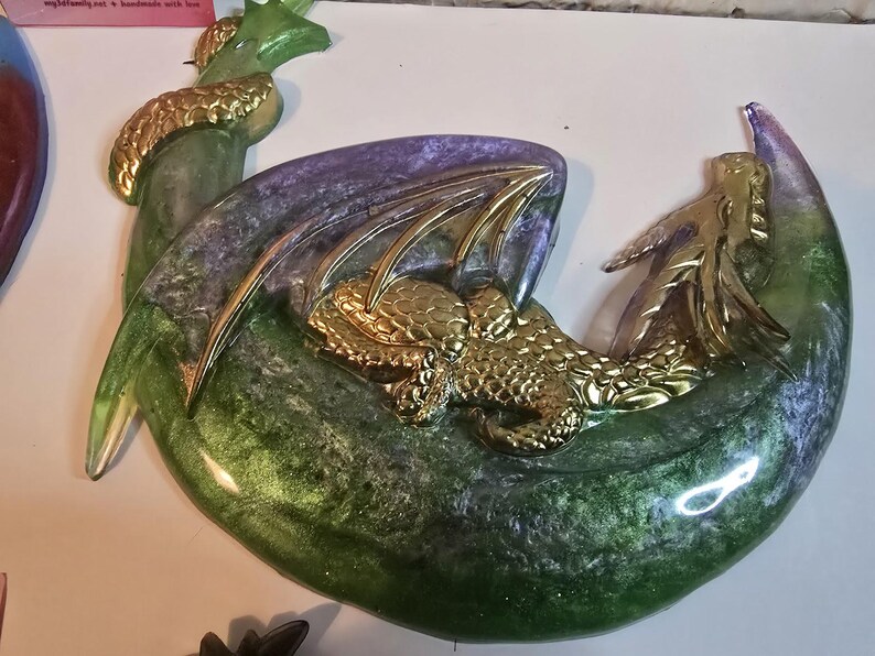 May include: A decorative dragon sculpture in shades of green, purple, and gold. The dragon is curled up, with gold-colored wings and accents. The base has a crescent moon shape. The sculpture is made of resin.