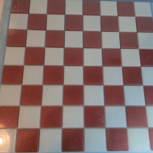 May include: A square chessboard with alternating red and white squares. The board is framed with a light blue border. The red squares have a glittery texture. A classic game board for chess or checkers.