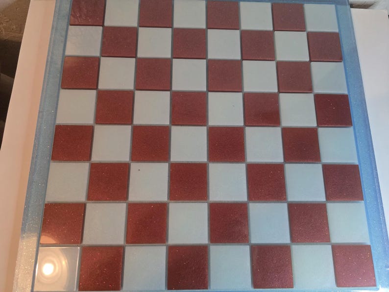 May include: A square chessboard with alternating red and white squares. The board is framed with a light blue border. The red squares have a glittery texture. A classic game board for chess or checkers.