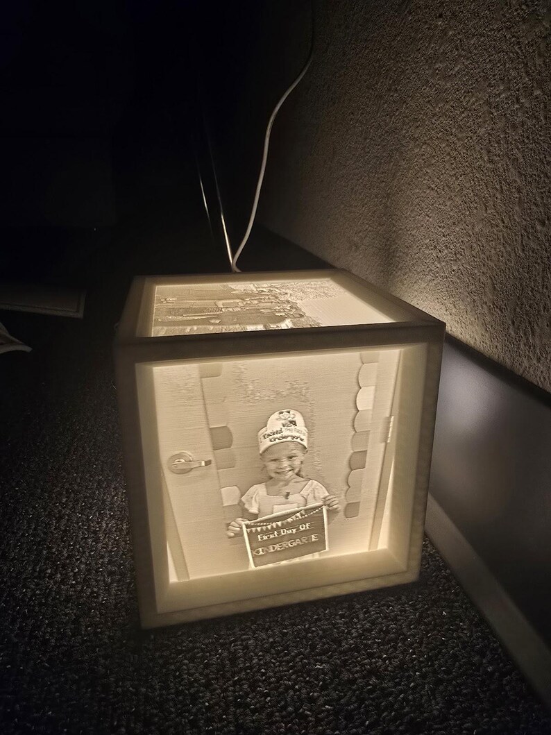 May include: A lit wooden box with a picture of a child holding a sign that says "First Day of Kindergarten". The box is illuminated from within, creating a warm glow. The image is a portrait of a child in a school setting.