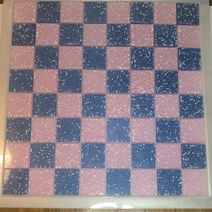 May include: A square, pink and blue checkerboard pattern with white speckles. The board is framed in a clear, plastic-like material. The squares alternate between pink and blue, creating a classic game board design.
