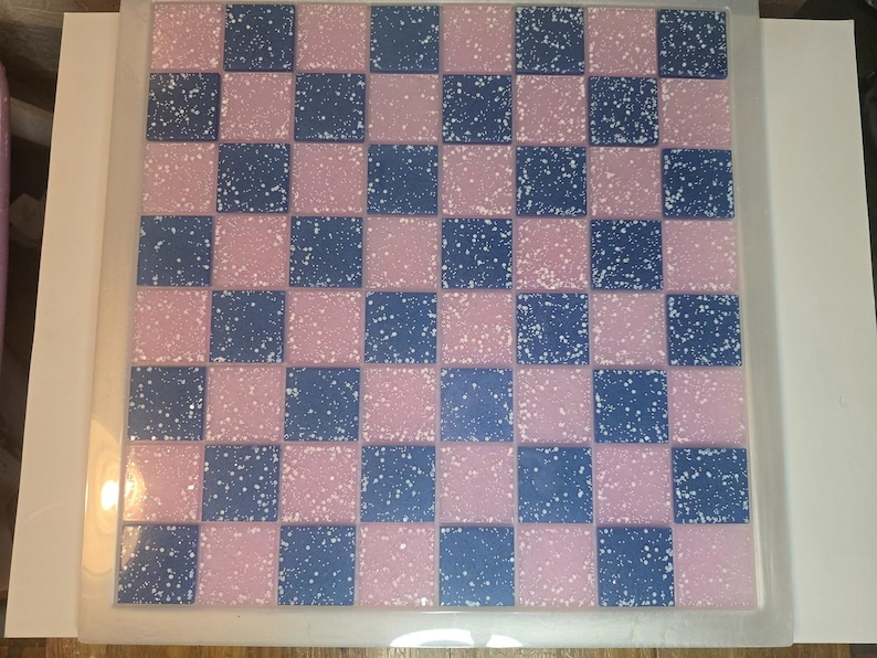 May include: A square, pink and blue checkerboard pattern with white speckles. The board is framed in a clear, plastic-like material. The squares alternate between pink and blue, creating a classic game board design.