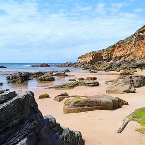 May include: A scenic coastal landscape featuring a sandy beach, rocky formations, and a cliffside. The water is calm, and the sky is blue with scattered clouds. The beach has a mix of sand and rocks, with some areas of green vegetation.