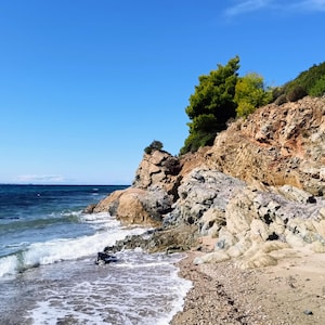 Könnte beinhalten: Eine malerische Küstenlandschaft mit Sandstrand, felsigen Klippen und dem blauen Ozean unter klarem Himmel. Wellen brechen am Ufer, mit grüner Vegetation und Bäumen auf den Klippen. Das Bild zeigt eine natürliche Landschaft.