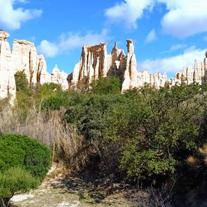 Könnte beinhalten: Panoramablick auf eine natürliche Felsformation mit hohen, erodierten Sandsteinsäulen. Üppige grüne Vegetation und Bäume umrahmen die Basis der Formationen. Der Himmel ist blau mit verstreuten Wolken.