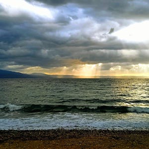 Könnte beinhalten: Eine malerische Meereslandschaft unter einem dramatischen Himmel. Dunkle, stürmische Wolken dominieren den oberen Teil, mit Sonnenstrahlen, die durchbrechen. Das Wasser ist dunkelgrün, mit kleinen Wellen am Ufer.