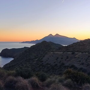 Könnte beinhalten: Landschaftsfotografie eines Küstengebirges bei Sonnenuntergang. Der Himmel wechselt von Orange zu Blau, mit Meer und Bergen als Silhouette. Der Vordergrund zeigt trockenes Gestrüpp.