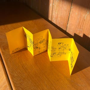 Op de afbeelding: Een felgeel, harmonica-achtig boekje met illustraties in zwarte inkt. Het boekje is open en toont verschillende panelen met kleine tekeningen en diagrammen. Het kunstwerk lijkt handgetekend en grillig, met een focus op eenvoudige vormen en lijnen.
