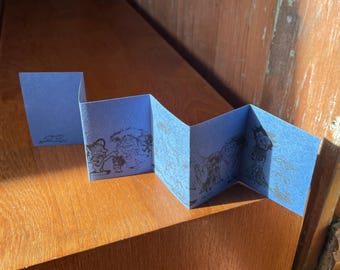 Puede incluir: Un folleto azul en acordeón con ilustraciones. El folleto está abierto, mostrando paneles con dibujos detallados en un color contrastante. El papel está doblado en zigzag, creando múltiples superficies de visualización.
