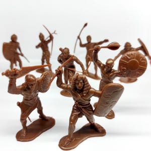 May include: A collection of brown plastic toy soldiers in various action poses. The figures are depicted with shields, spears, and bows, suggesting a historical or fantasy theme. The toys are on a white background.