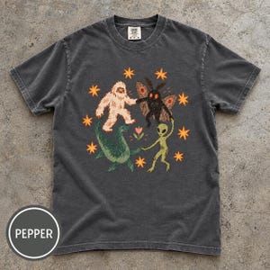 May include: A charcoal gray short-sleeve t-shirt featuring a graphic print of a white yeti, a black mothman, a green sea monster, and a green alien holding hands in a circle around a small flower, surrounded by orange stars. The label reads Comfort Colors.