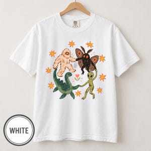 Può includere: Una t-shirt bianca a maniche corte con un'illustrazione grafica di uno yeti beige, una creatura nera simile a una falena, un dinosauro verde e un alieno verde che si tengono per mano in cerchio attorno a un piccolo fiore.