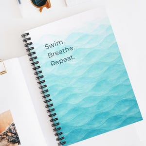 May include: Spiral-bound notebook with a blue watercolor wave design. The notebook features the text "Swim. Breathe. Repeat." in a teal font. The notebook is open on a white surface, with sunglasses and a notepad in the background.